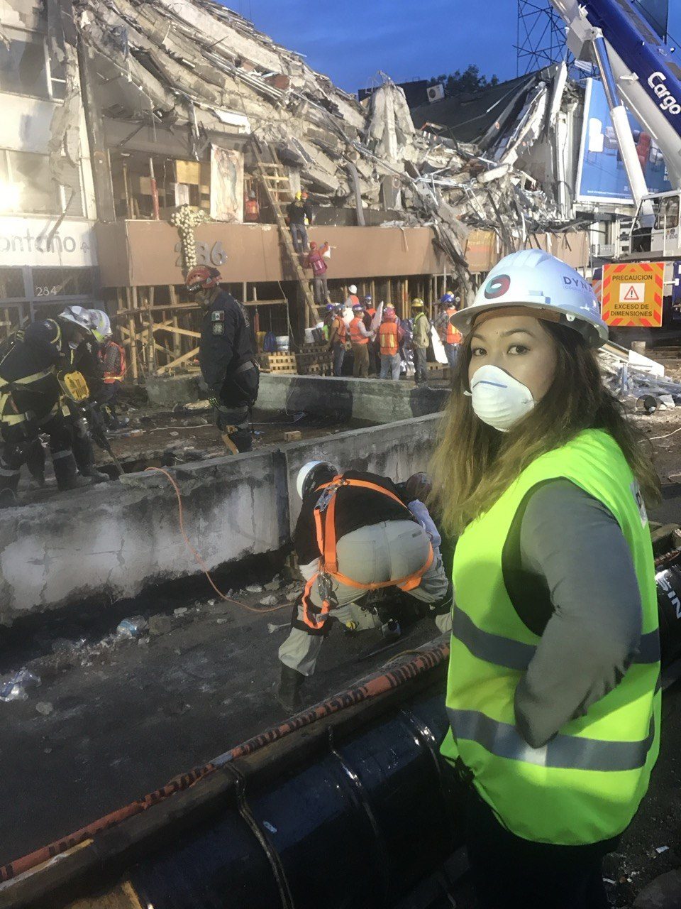 Dynamis engineer in a DYNA hi-vis vest, hardhat and mask before a collapsed building in Mexico City, 2017; a rescuer rappels down the structure.